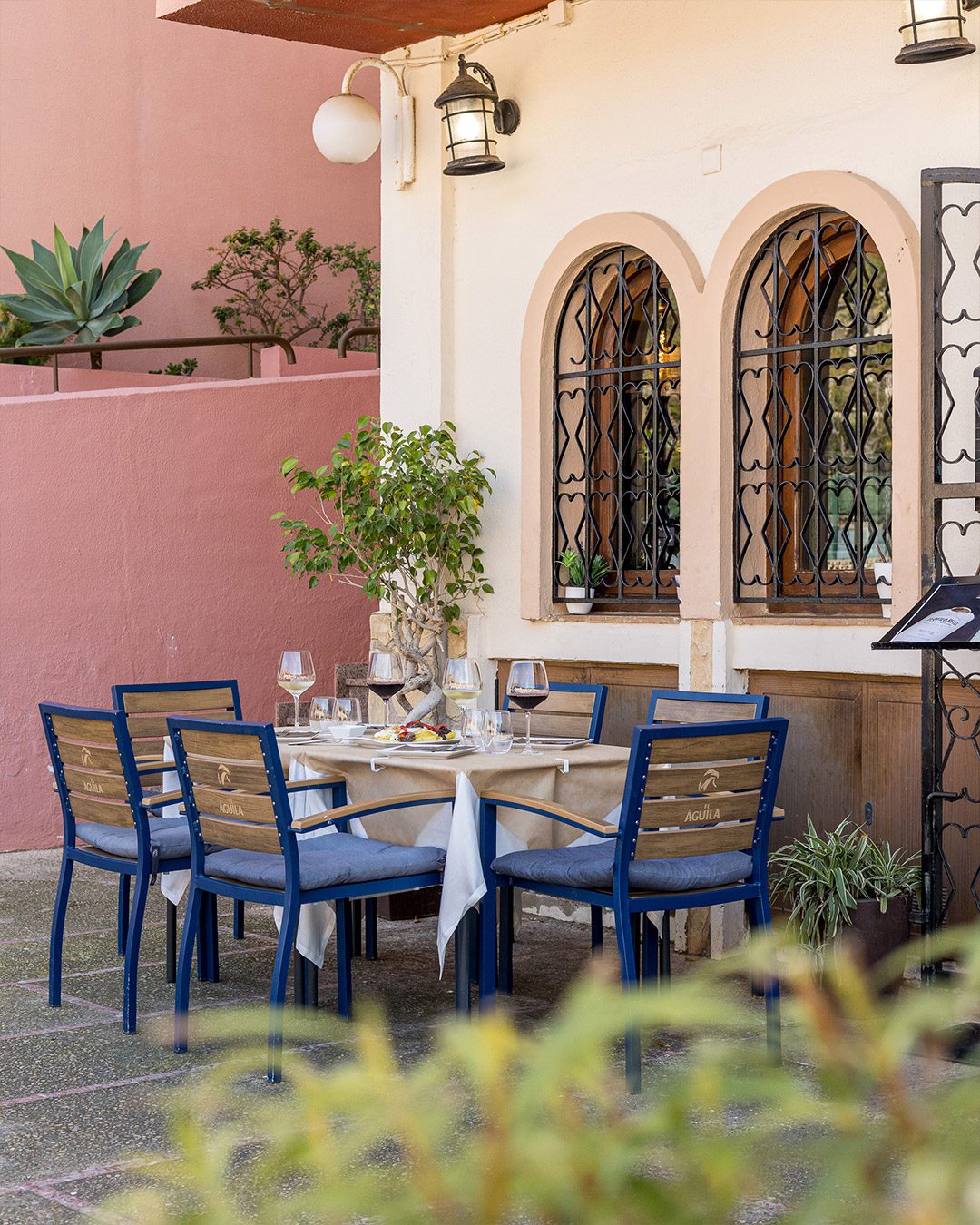 Una mesa preparada con platos y copas de vino en la terraza exterior de la Pizzería Crepería Stromboli en Altea. Destacan las sillas azules con lamas de madera dispuestas frente a una fachada blanca con ventanas de arco rústicas y rejas de forja.